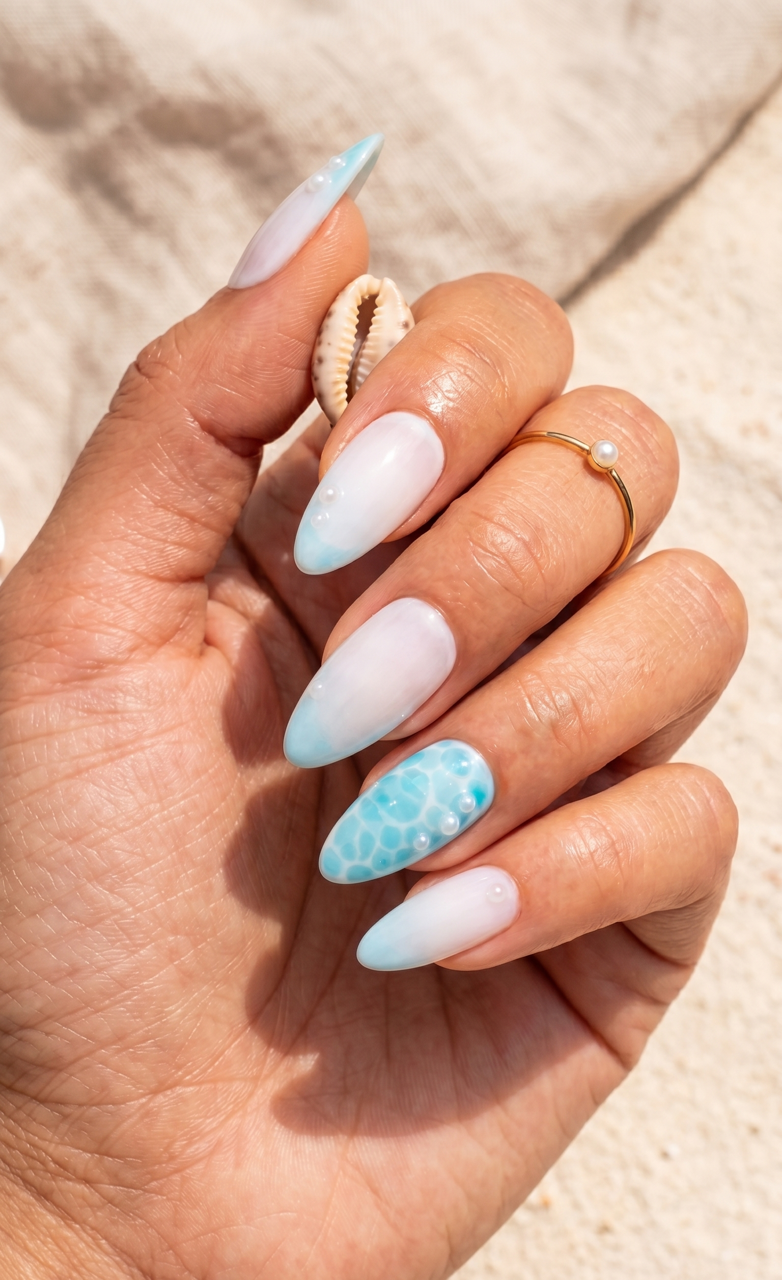 White-to-sky-blue ombre stiletto nails, accent nail with turquoise water ripple effect and pearls, beach background